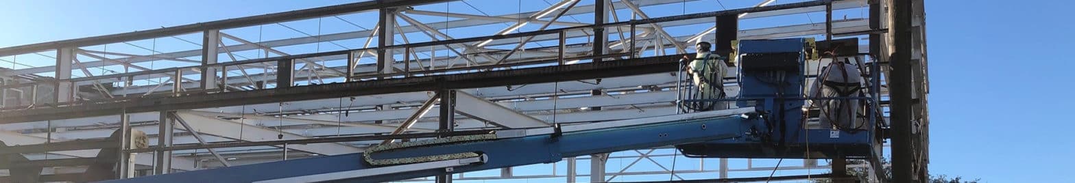Two workers in protective gear stand on a blue aerial lift platform, inspecting or working on the framework of a large metal structure under clear blue skies, showcasing the precision required by an industrial hygiene services team.