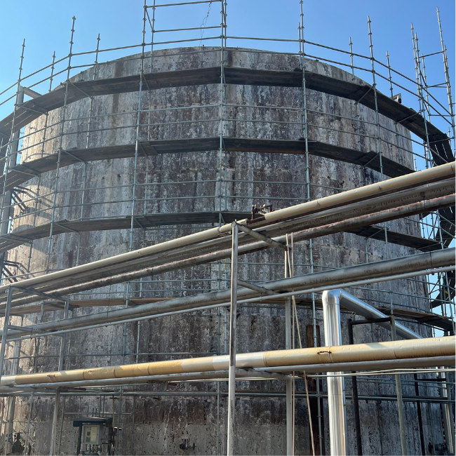 A large, cylindrical industrial structure with scaffolding around it. Multiple pipes are visible in the foreground, and the sky is clear. A lead-based paint inspection is underway to ensure safety compliance.