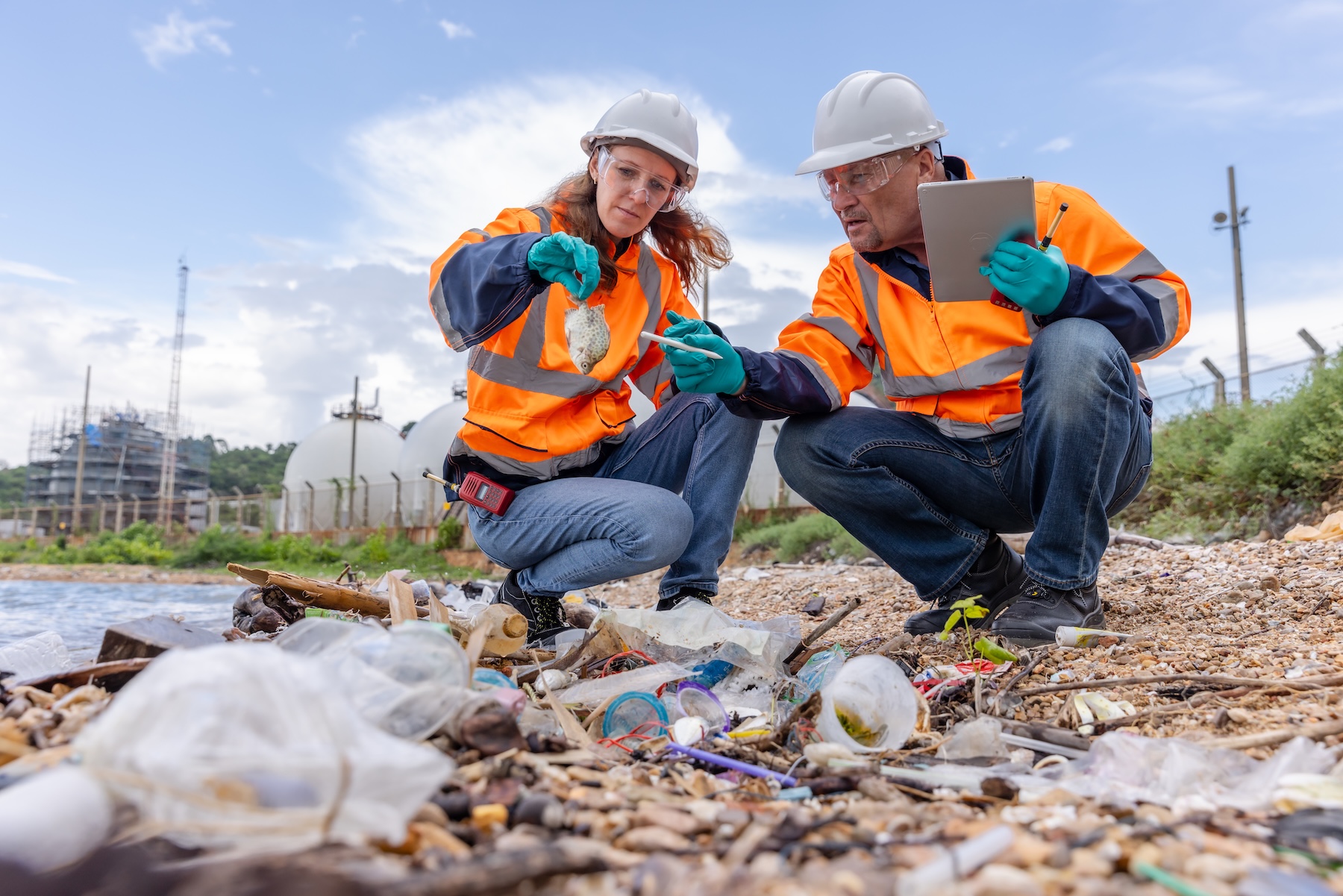 Two workers in safety gear examine and document solid waste permitting issues on a rocky shoreline near an industrial area.
