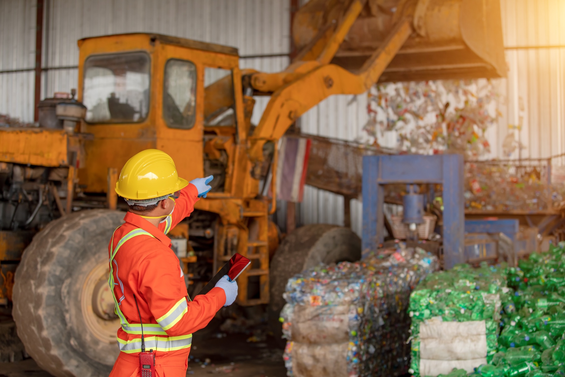 Worker in a safety helmet and vest points toward machinery and compacted plastic bottles, ensuring Solid Waste Permitting compliance in a recycling facility.